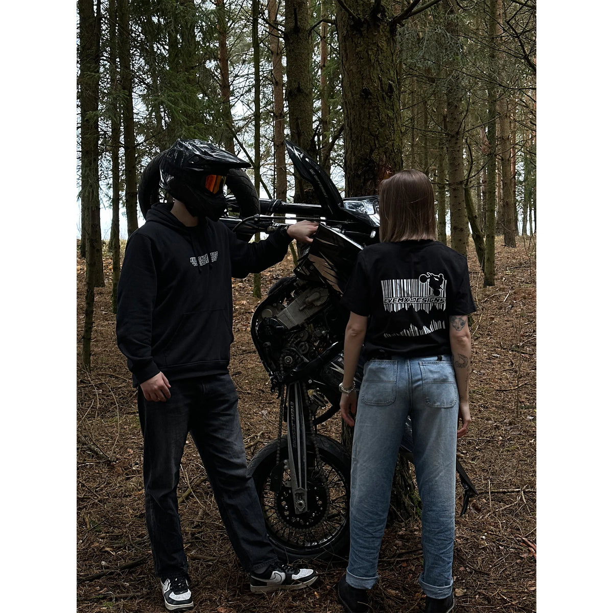 A person in a black hoodie and helmet stands next to a black motorcycle in a forest, while another person in a black graphic T-shirt and light blue jeans stands facing them. The scene is set in a wooded area with tall trees and brown forest floor.