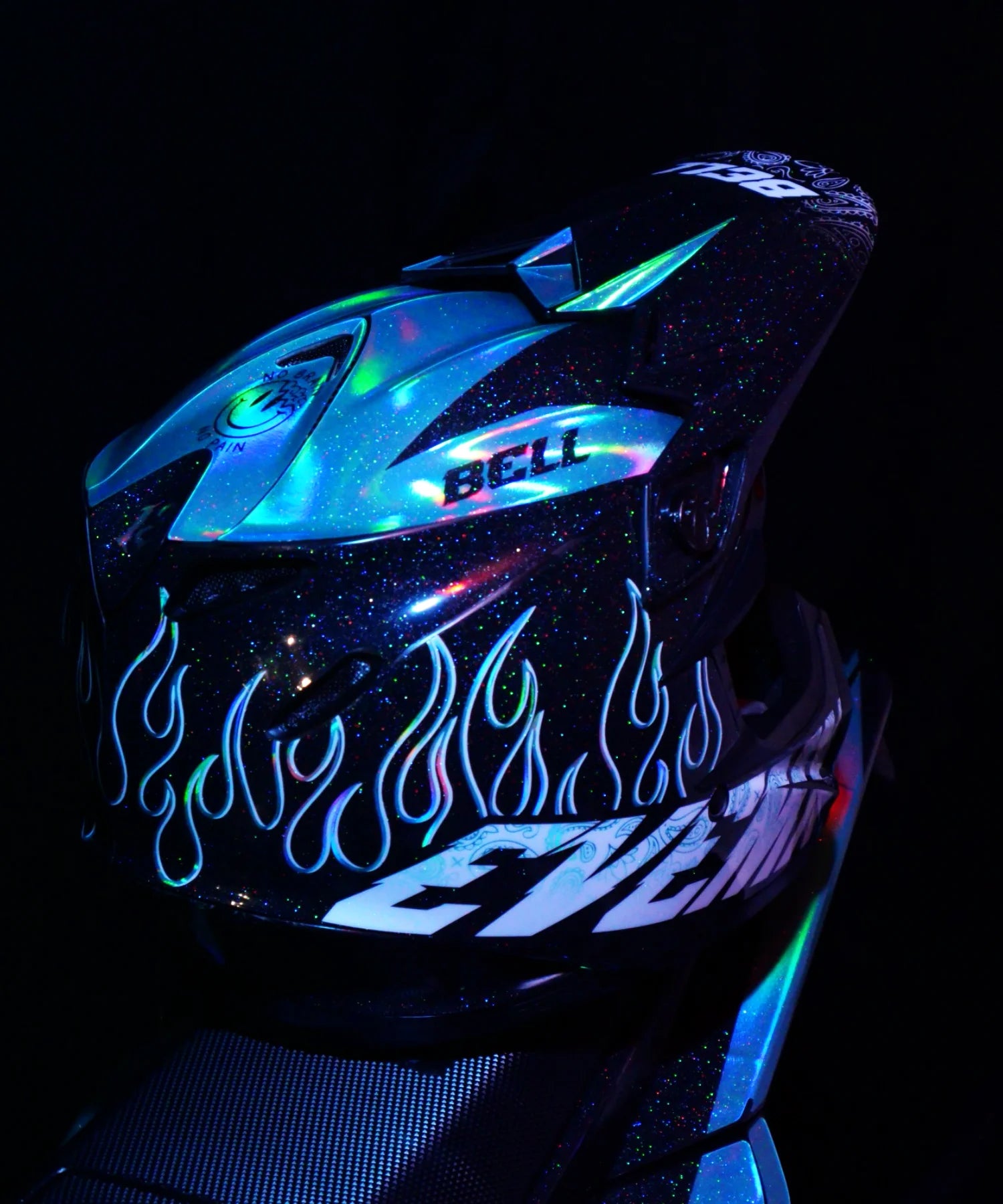 A close-up of a Bell full-face motorcycle helmet with a full holografic chrome, blue and white flame graphics, and a clear visor, shown against a dark background.