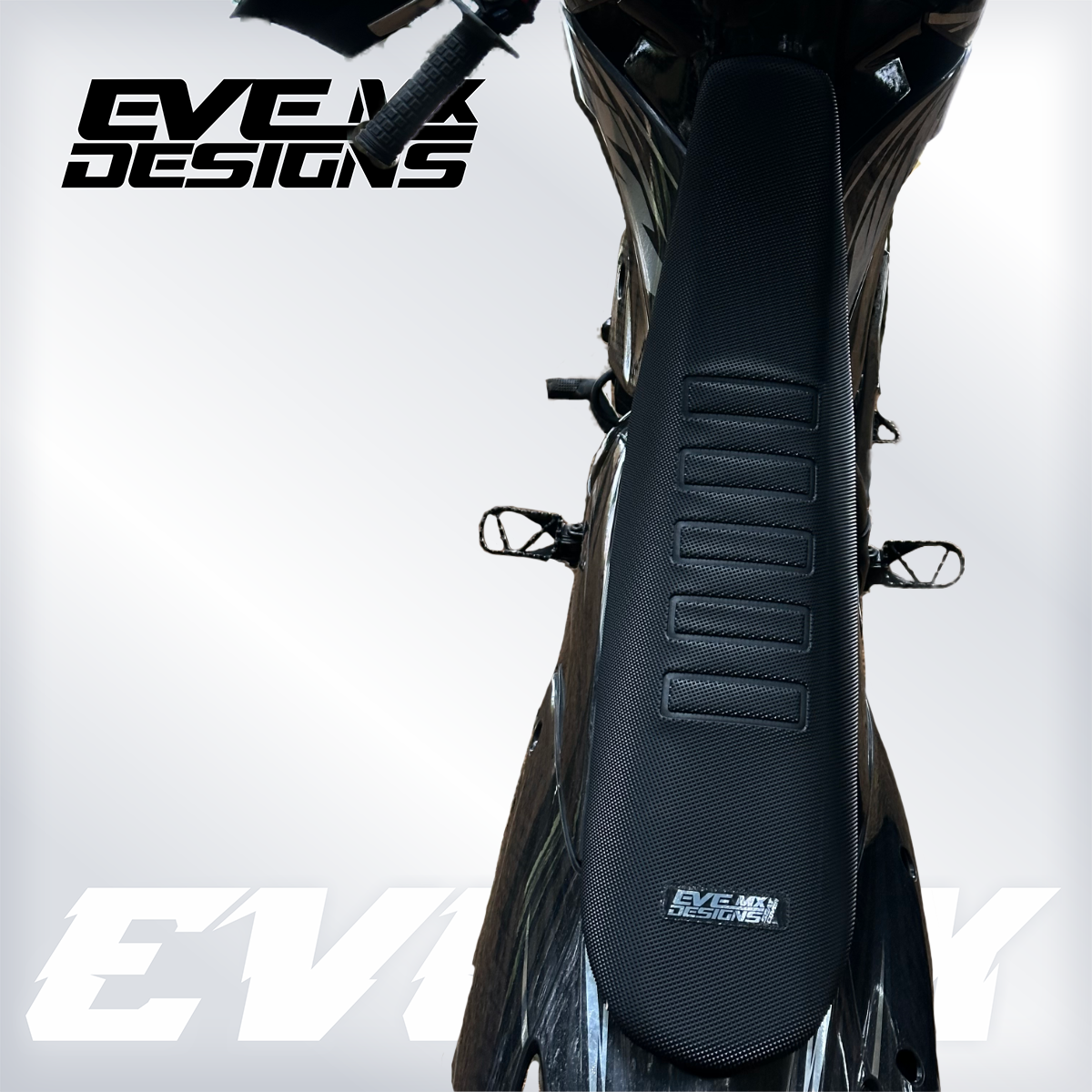 Close-up of a motorcycle seat with 'EVE MX DESIGNS' branding on a light gray background.